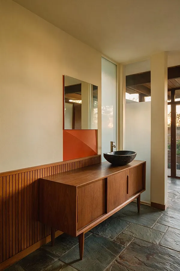 A mid-century modern powder room with a long walnut vanity on tapered legs, topped by a black vessel sink and brushed metal faucet. A rectangular mirror with a burnt orange backing hangs above vertical wood slats, while pale beige walls, a large frosted window, and a slate stone floor create a warm, restrained atmosphere.