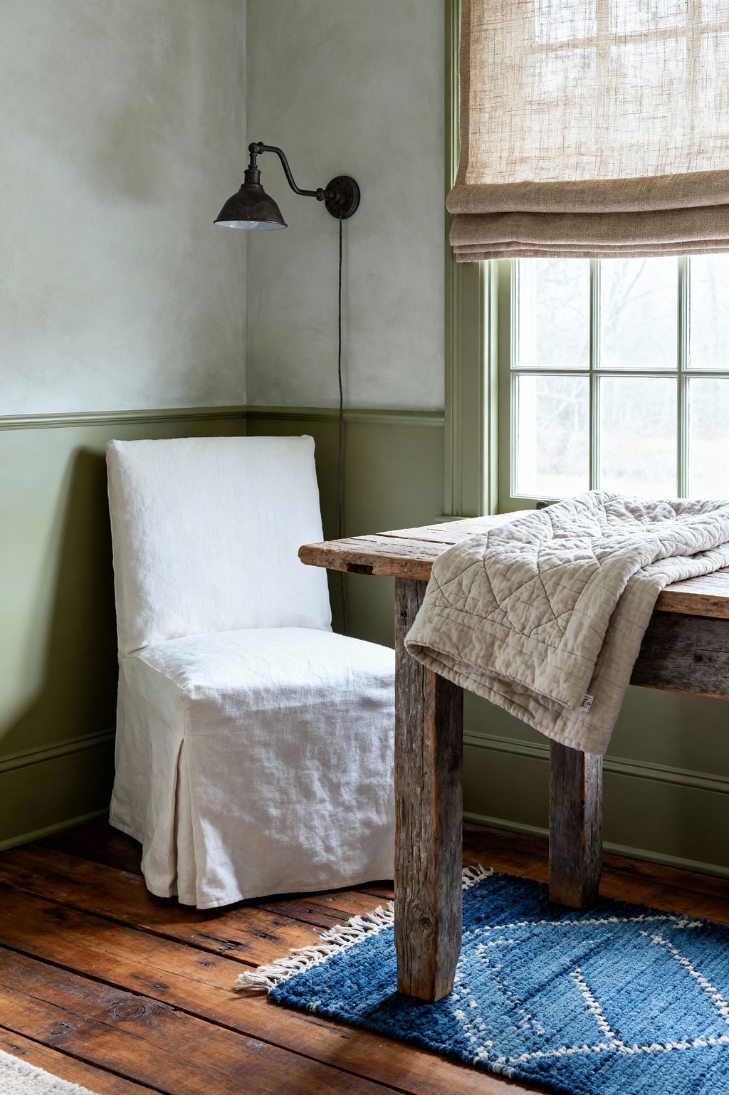 Farmhouse living room corner featuring a rustic reclaimed-wood table by a window with sage green trim and a natural linen Roman shade. A white linen slipcovered chair sits on wide-plank pine floors with a small indigo rug; an oatmeal quilt is draped over the tabletop, and a vintage blackened-iron wall sconce hangs above pale plaster walls, all softly lit by daylight.