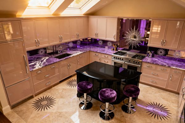 An L-shaped kitchen features champagne-gloss cabinets with chrome pulls, purple marble countertops, and a purple-tinted mirrored backsplash decorated with repeating sunburst designs, including a large starburst behind a stainless-steel range. A small black curved island sits at center with three chrome-base stools upholstered in plush purple velvet, set on cream marble floors with starburst inlays, while skylights flood the space with warm natural light.