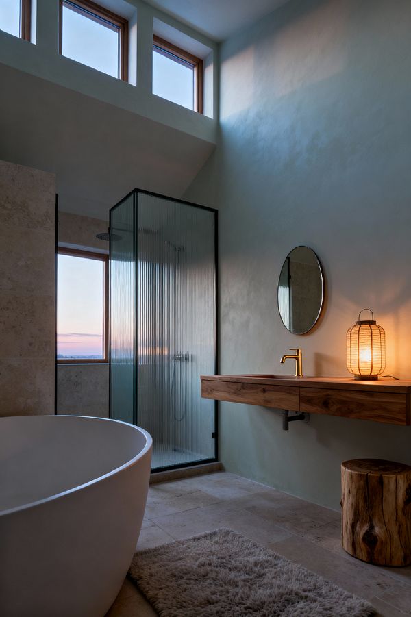 A serene bathroom features a freestanding matte white tub, a black-framed shower with reeded glass, and a floating solid-wood vanity topped with a round mirror and brass faucet. Warm light from a woven lantern and high clerestory windows illuminates beige limestone floors and pale sage plaster walls, with a log stool and soft rug adding natural texture and a calm, Scandinavian feel.