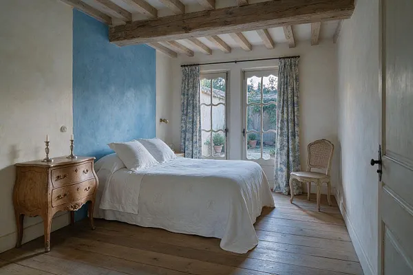A cozy French Country guest bedroom with exposed wood ceiling beams, wide-plank wooden floors, and plaster walls. A bed dressed in white bedding sits beside a distressed wood nightstand, while floral curtains frame French doors that open to a small outdoor courtyard.