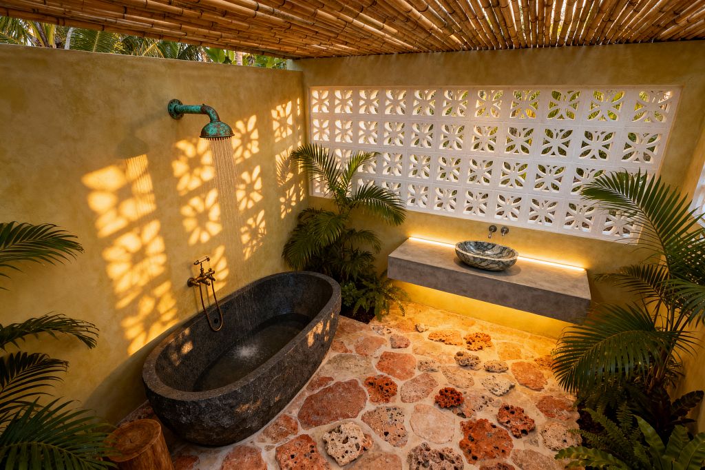 Open-air tropical bathroom with a bamboo slat roof, sun-washed ochre plaster walls, and a white breeze-block screen casting floral shadows. A dark basalt soaking tub sits beneath a patinated copper rain shower; a cast-concrete floating vanity with warm underlighting holds a stone vessel sink, while coral stone pavers, potted palms, and ferns complete the space.