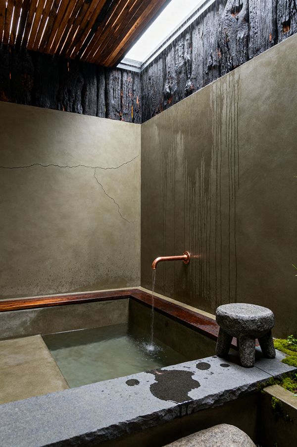 An intimate bathroom with a deep concrete soaking tub framed by dark-stained wood and a copper spout trickling water. Smooth lime-plaster walls show a hairline crack and vertical water marks. Light enters from a skylight screened by timber slats and surrounded by blackened, charred wood. A rounded granite stool rests near a rough stone ledge with wet splashes and a patch of moss. The scene is minimal, tactile, and quietly lit.