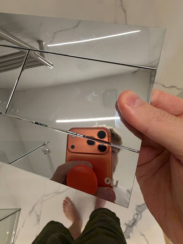 Upside-down installed toilet flush button held in hand with reflective surface showing a person's reflection taking a photo with an orange phone.