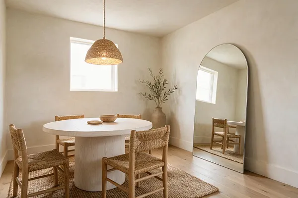 A serene dining room with a round white pedestal table surrounded by woven natural wood chairs, set on a textured jute rug. A woven pendant light hangs overhead, and a tall arched mirror leans against the wall beside a large ceramic vase with greenery.