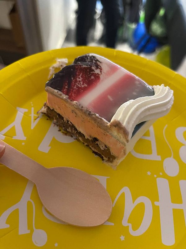 A slice of colorful cake is presented on a yellow plate with a wooden spoon.