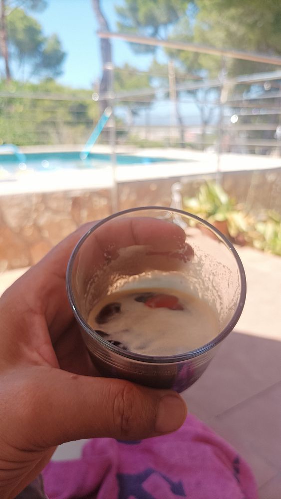A hand holds a glass of iced espresso with a pool and greenery in the background.
