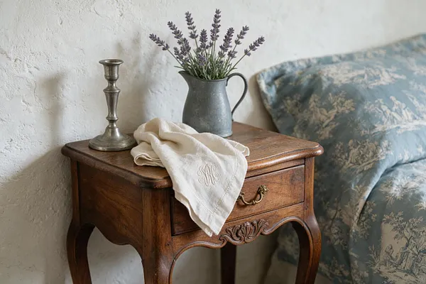A small vintage wooden nightstand with curved legs stands beside a bed with blue toile bedding. On top are a brass candlestick, a gray metal pitcher holding lavender, and a folded cream monogrammed linen cloth, all set against a textured white wall.