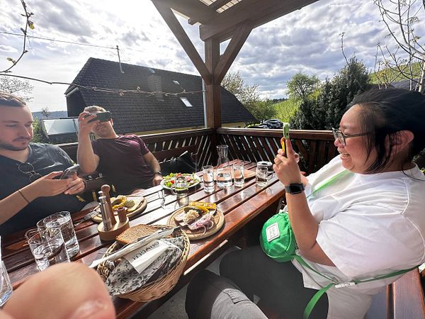 A group of three people enjoys a meal on a wooden terrace, capturing the moment with their phones.