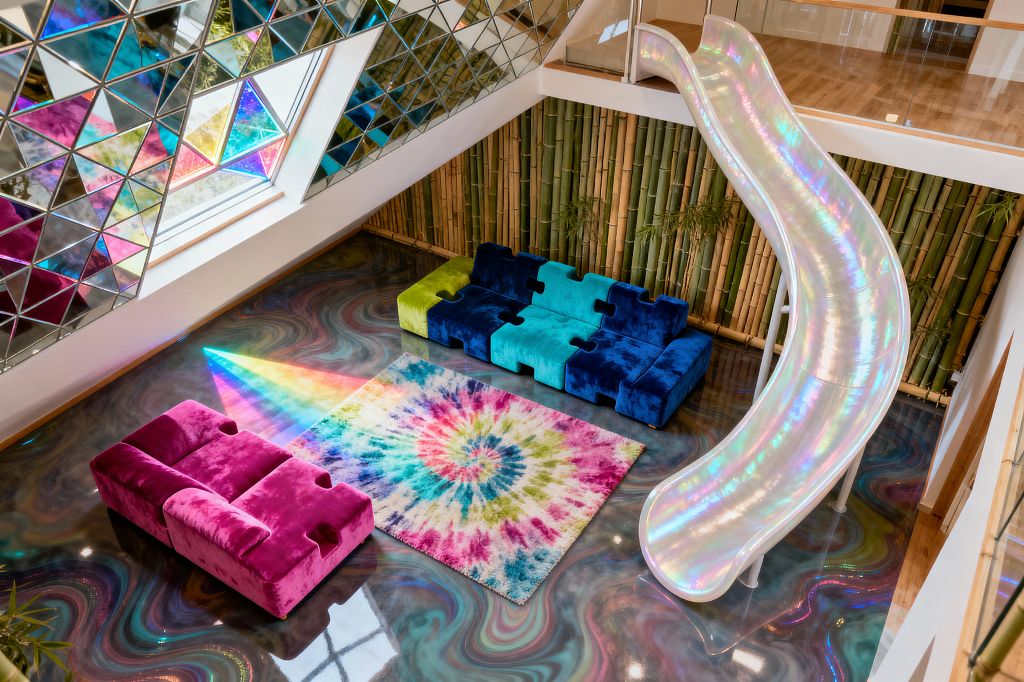 A double-height kids’ playroom features an iridescent curving slide descending from a mezzanine with a glass balustrade onto a glossy, marbled epoxy floor. A tie-dye rainbow rug sits between low modular velvet sofas—one fuchsia, one in blue, teal, and lime—while a wall of vertical bamboo adds natural texture and a triangular mirrored-and-colored glass window scatters prismatic light across the room.