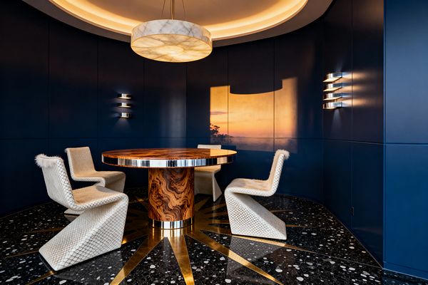 An Art Deco dining room with curved navy lacquered wall panels surrounds a round burled-wood pedestal table edged in chrome. Four sculptural ivory chairs with diamond-quilted upholstery and soft shearling seats encircle the table. A large alabaster drum pendant and recessed cove lighting cast a warm glow, while three-tier chrome sconces punctuate the walls. Underfoot, black terrazzo flooring with white aggregate features a brass starburst inlay radiating from the table’s base.