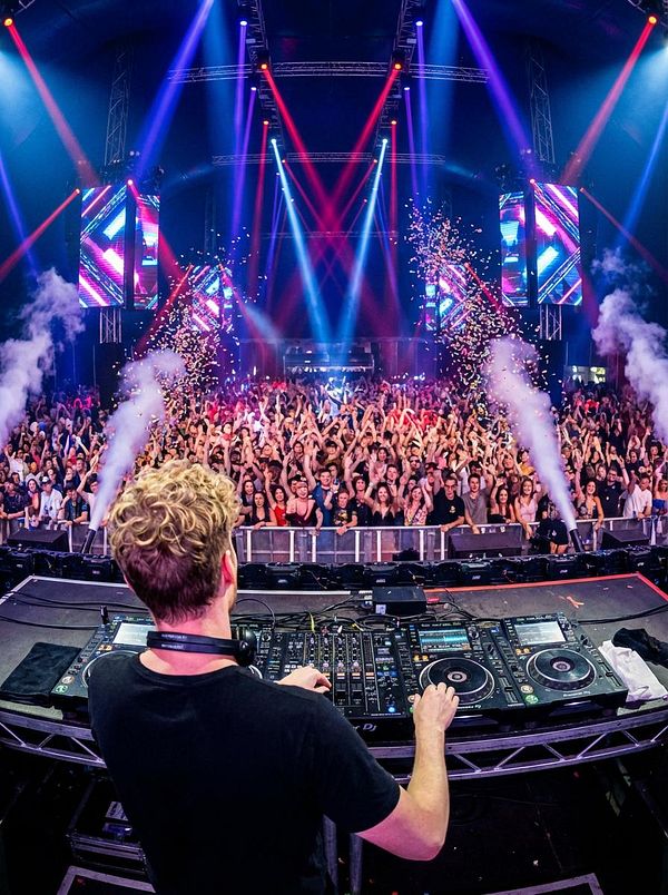 A DJ performs in front of a large, energetic crowd at a vibrant music festival.