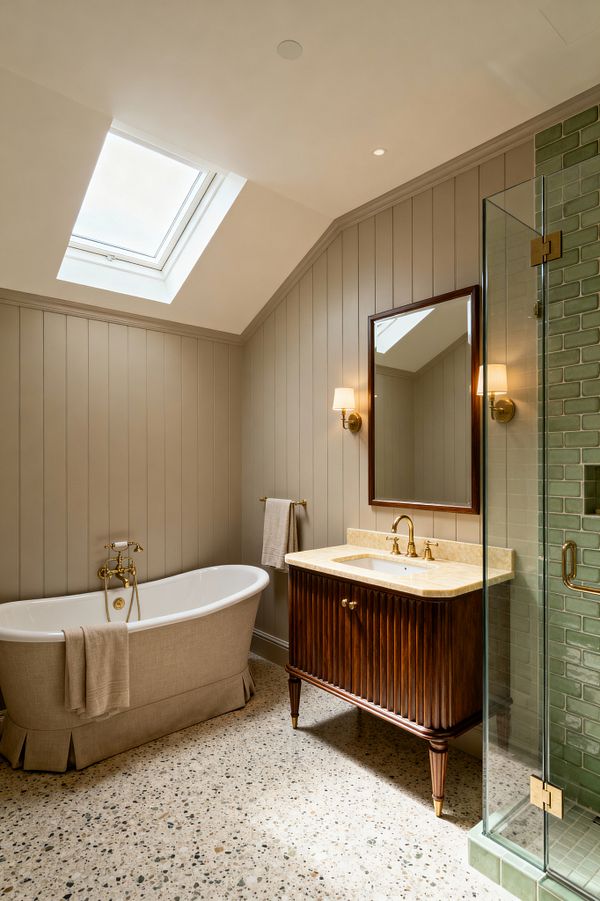 A transitional bathroom with a sloped ceiling and skylight features vertical greige wall paneling and a terrazzo floor. A freestanding white tub wrapped in a tailored linen skirt sits beside a brass tub filler. Opposite is a fluted dark-wood vanity with a cream stone countertop, undermount sink, and brass faucet, topped by a wood-framed mirror and brass sconces with white shades. A glass shower reveals glossy sage-green subway tiles and brass hardware.