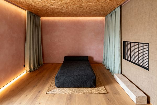 A minimalist Scandinavian media room centers on a low charcoal wool daybed set on a woven jute rug over warm oak floors, framed by blush plaster walls and a natural OSB wood ceiling with hidden perimeter lighting. Soft sage linen curtains pool at both sides, while a textured beige wall with a horizontal black metal grille and a slim travertine block complete the calm, tactile space.
