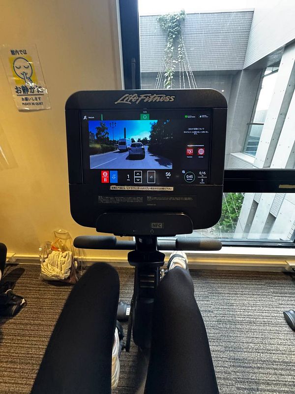 A person is using a stationary bike in a gym, with a view of a screen displaying a virtual cycling route.