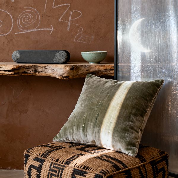 Close-up of a bohemian media room with a warm terracotta plaster wall marked by chalk doodles, a live-edge driftwood shelf holding a charcoal speaker and a small green ceramic bowl, and a woven seagrass ottoman with a black geometric pattern topped by an olive velvet pillow with a pale stripe. At right, a vertical reeded glass panel glows with a crescent moon projection, adding soft, shimmering light to the tactile, earth-toned scene.