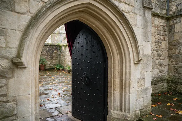 A Gothic stone exterior features a pointed arched doorway framed in pale carved stone, with a heavy black wooden door studded with iron rivets and a central ring handle. The wet stone paving, mossy walls, and dark drapery inside the opening create a moody medieval atmosphere.