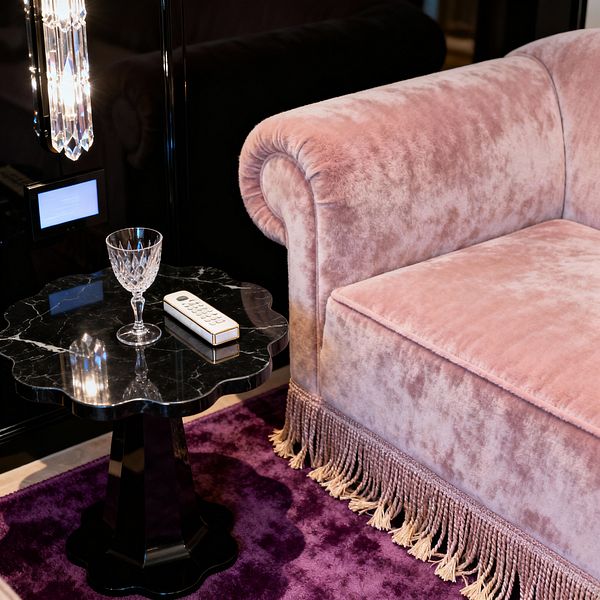 A close-up of a vintage glam media room shows a blush pink velvet sofa with a rolled arm and bullion fringe along the base. Next to it, a scalloped black marble side table holds a cut-crystal goblet and a white remote with gold trim. A deep purple rug anchors the scene, while glossy black wall panels reflect light from a faceted crystal sconce, creating a polished, luxurious atmosphere.
