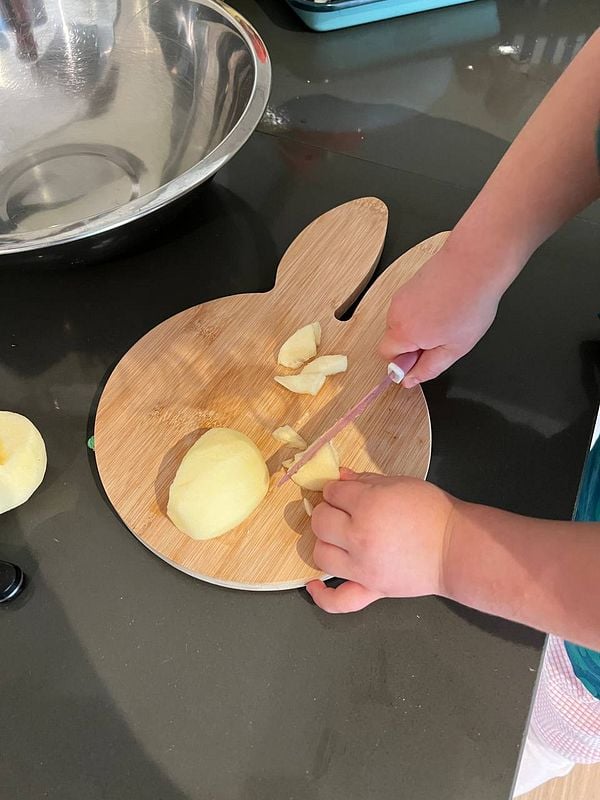 A child is peeling an apple on a bunny-shaped cutting board in a kitchen setting.