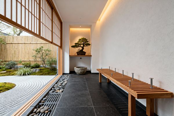A minimalist mudroom opens to a Zen garden through large shoji screens with wooden lattice and translucent paper. Inside are charcoal stone tiles, a pebble-and-drain channel along the threshold, and a long slatted wood bench topped with short stainless pegs; opposite, a recessed alcove displays a bonsai on a floating shelf above a rough stone basin, all lit by warm cove lighting, with white plaster walls and a bamboo fence outside.
