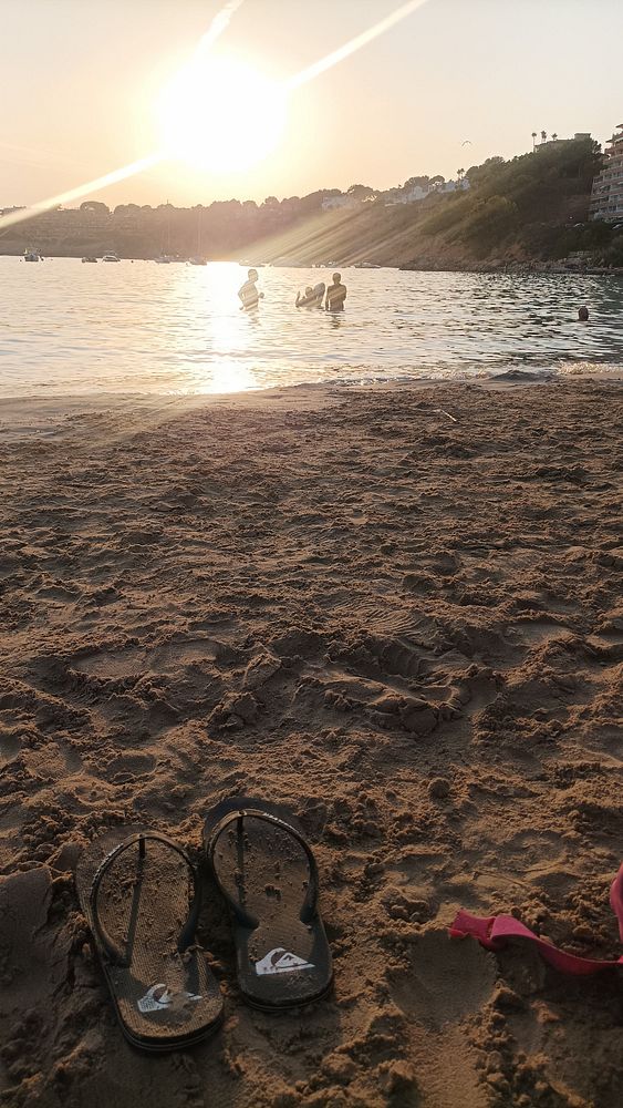 A serene beach scene at sunset with flip-flops in the foreground and people enjoying the water.