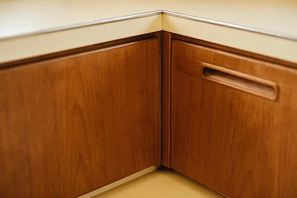 Close-up of a mid-century modern corner cabinet with warm walnut wood grain and a cream laminate top. One cabinet door features a recessed handle, and the clean angled joinery emphasizes the geometric lines typical of the style.