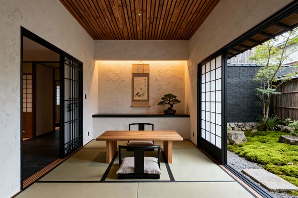 A Japanese Zen-style home office features a tatami floor with a low solid-wood desk and two floor chairs, set before a backlit alcove displaying a hanging scroll and a bonsai. Textured plaster walls, a slatted wood ceiling, and black-framed openings lead to shoji sliding doors that open to a small moss garden with stones and a tree, filling the minimalist space with soft natural light for quiet work.