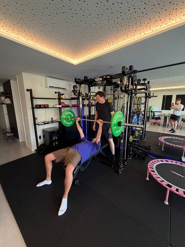 Three men posing at the gym, holding resistance bands, in front of a mirrored fitness studio wall.