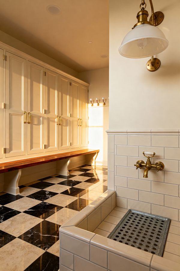 A traditional mudroom features cream paneled built-in cabinets with brass hardware and a long solid wood bench on decorative brackets. The floor is a polished black-and-beige marble checkerboard. At right, a small tiled utility sink with glossy cream subway tiles, a galvanized grate, and an unlacquered brass faucet sits below a brass-and-opal glass sconce, while a peg rail with white knobs catches warm late-afternoon light.