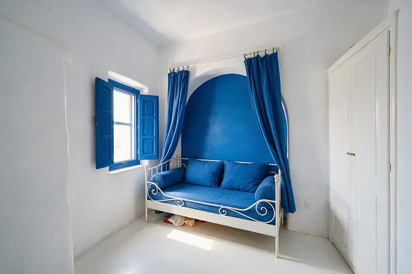A bright kids' room with white plaster walls, a vivid blue alcove, matching blue curtains, and an iron daybed with blue cushions. A blue shuttered window brings in natural light, while a white double-door closet keeps the room simple and airy.