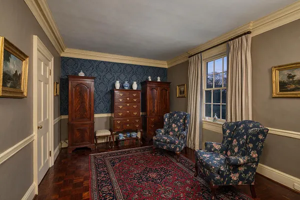 A traditional closet or dressing room with two floral wingback chairs, a red patterned area rug, and polished dark wood furniture including a chest of drawers flanked by tall armoires. The room has taupe walls with cream trim, blue damask wallpaper on the far wall, a large window with ivory curtains, and framed landscape artwork.