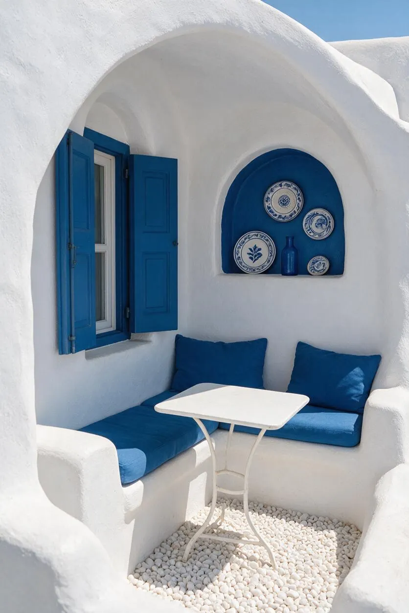A small Mediterranean-style patio with smooth white stucco walls, rounded built-in seating, and bright blue cushions and window shutters. A niche displays blue-and-white ceramic plates above a small white table set over a pebble floor under natural sunlight.