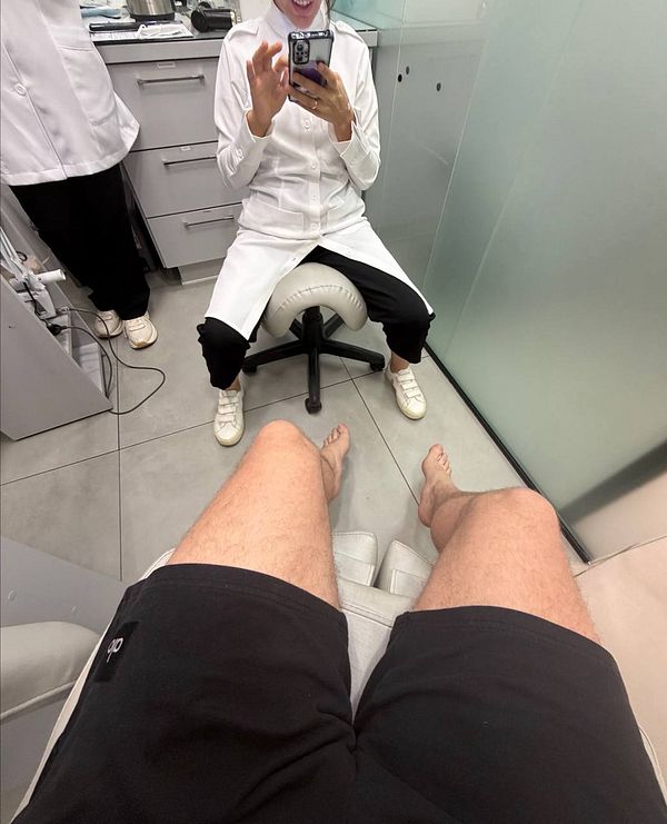 A woman with long black braided hair wearing a black top and purple skirt stands in a green garden holding a leash attached to a dog.