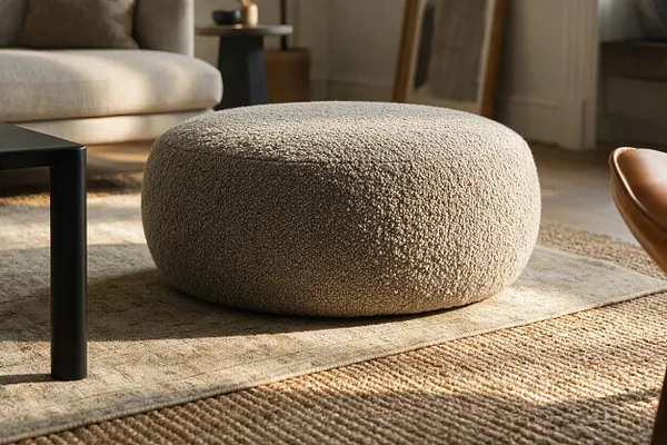 A large round ottoman upholstered in a textured bouclé fabric sits on a layered neutral rug in a sunlit living room. Nearby are a cream sofa, a black side table, and a tan leather chair, creating a calm transitional setting.