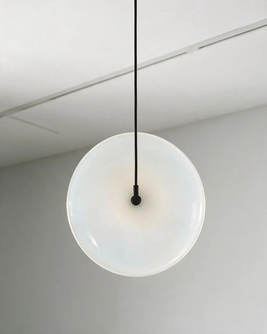 A minimalist pendant light hangs from the ceiling on a slim black cord, centered above a large frosted white circular glass shade. The clean gray wall and ceiling create a soft, airy backdrop that emphasizes the light's sculptural simplicity.