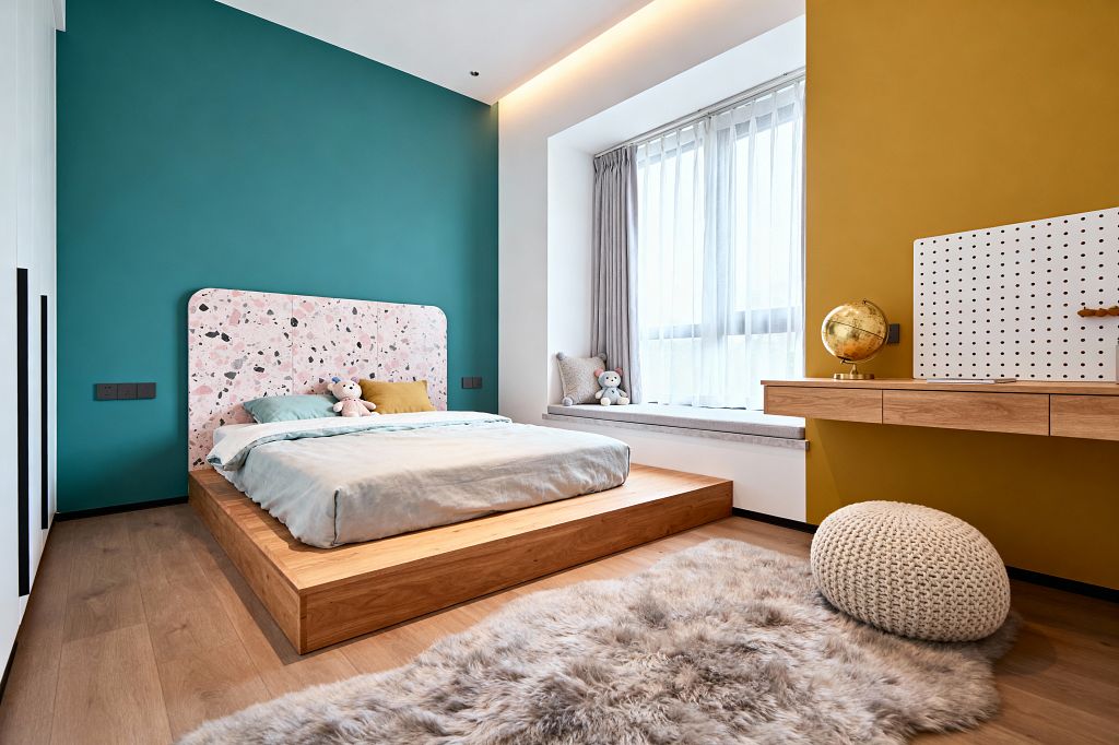 A modern children’s bedroom with a low light-oak platform bed, mint bedding, and a rounded pink terrazzo-pattern headboard set against a teal wall. A mustard accent wall features a floating wood desk with a white pegboard and gold globe, while a large window with sheer curtains and a cushioned bench brightens the space; a knitted cream pouf and fluffy rug soften the oak floor, and concealed cove lighting adds a warm glow.
