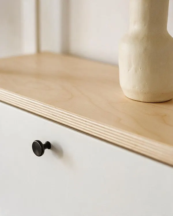 A close-up of a Scandinavian-style cabinet with a light birch plywood top, visible layered edge detail, and a matte white front panel. A small round black knob adds contrast, and a pale ceramic vase sits on the wood surface against a soft neutral background.