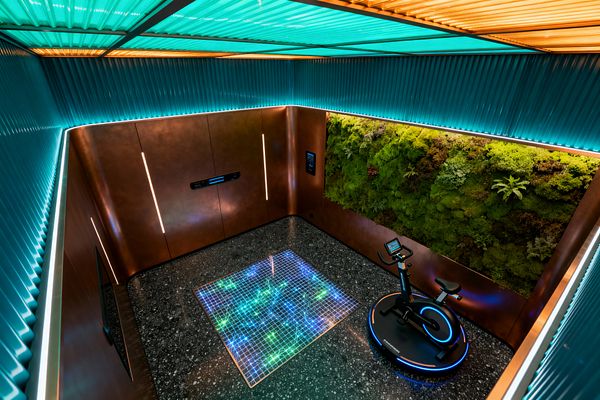 A compact, high-tech home gym features bronze metal walls inset with vertical light strips, a floor of dark speckled terrazzo, and a square LED grid set into the floor that glows in teal and green. One wall is a lush moss garden framed by copper, while a black stationary bike sits on a circular base lit with neon blue; ribbed turquoise panels and cove lighting wash the ceiling in aqua and amber.
