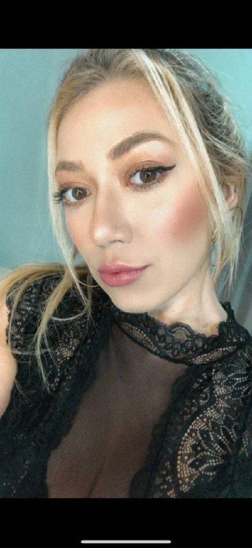 A close-up selfie of a woman with long, light hair and makeup, wearing a black lace top.