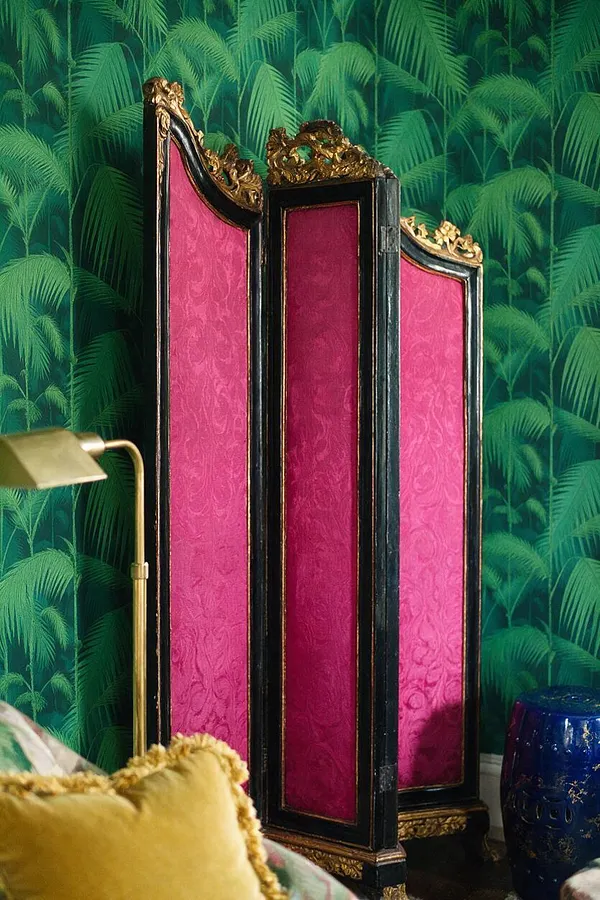 A tall three-panel folding screen stands against tropical palm-print wallpaper. The screen has black lacquered frames with ornate gold carvings and vivid hot pink velvet-like panels, creating a dramatic maximalist focal point.
