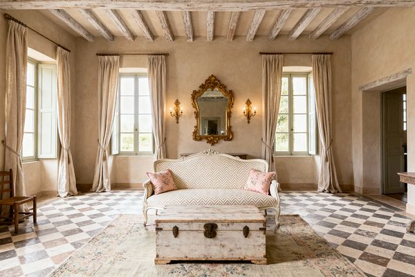 A sunlit French Country living room centers on a cream Louis XV settee in herringbone upholstery, flanked by blush floral pillows, facing a weathered white trunk on a faded floral rug. Behind, a gilded rococo mirror and pair of candle-style sconces hang on limewashed plaster walls between tall windows with sage-painted casements and linen drapery. Whitewashed ceiling beams, a gray-and-ivory stone checkerboard floor, and a rustic side chair add patina and symmetry to the airy, elegant space.