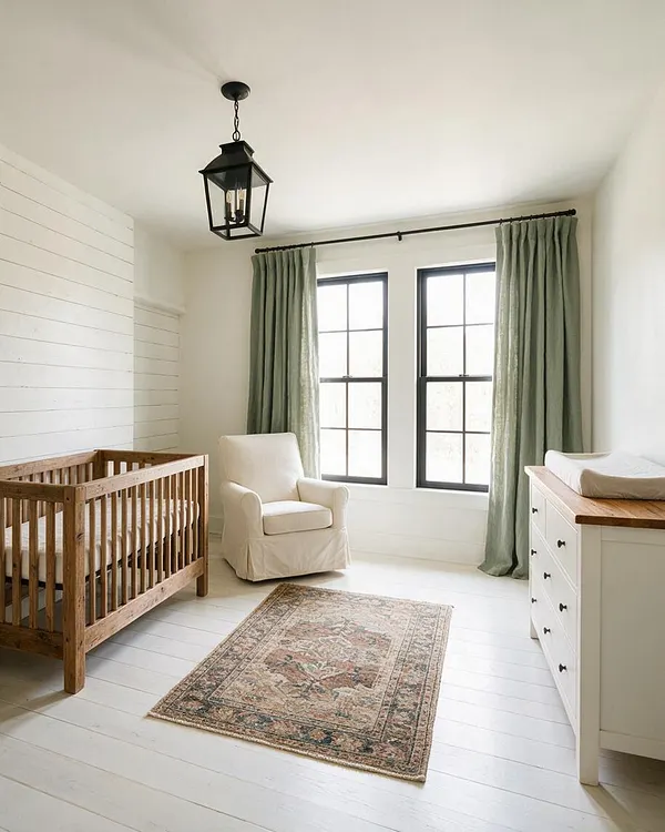 A bright farmhouse nursery with white shiplap walls, black-trimmed windows, and sage green curtains. The room includes a natural wood crib, a cream upholstered armchair, a white dresser with a wood top, and a muted vintage-style rug under a black lantern ceiling light.