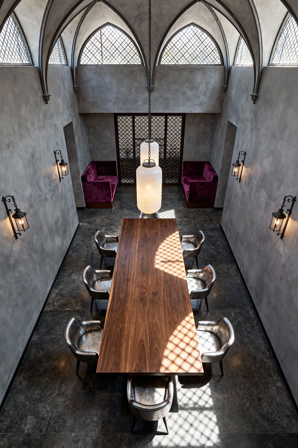 A Gothic-style dining room with a soaring ceiling of ribbed, pointed vaults and high leaded-glass windows. A long walnut table sits at center, surrounded by silver-toned leather chairs, with wrought-iron lantern sconces on gray limewashed plaster walls and a dark stone floor. Two chain-hung cylindrical alabaster pendants hang above, and plum velvet banquettes sit in rear niches beside a decorative iron screen, with patterned light falling across the space.