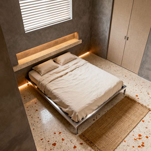 Minimalist bedroom featuring a low, metal-framed platform bed dressed in beige linen against smooth gray microcement walls. A long recessed niche forms a terrazzo ledge above the headboard, illuminated by warm LED strips that also trace the wall base; white terrazzo flooring with terracotta chips and a narrow jute runner ground the scene, with flat-panel beige wardrobe doors to the right and a window with horizontal blinds filtering daylight.