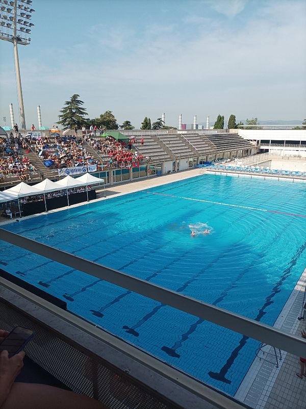 A synchronized swimming competition is taking place in a large outdoor pool with spectators in the stands.