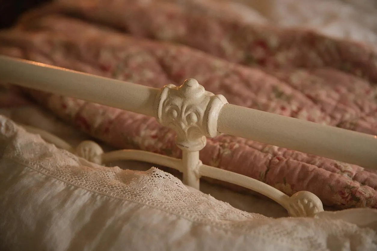 Close-up of a vintage white metal bed frame with ornate floral detailing on the headboard or footboard, layered with soft pink floral quilts and a cream lace-trimmed coverlet. The bedding creates a warm, romantic Cottagecore atmosphere with muted, nostalgic tones.