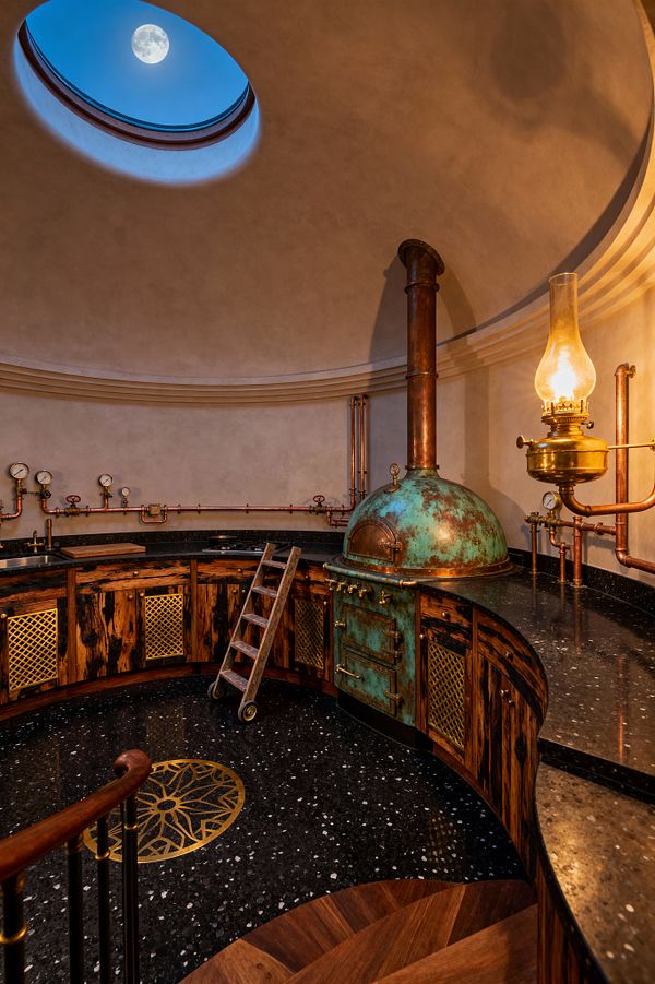 A circular steampunk kitchen set inside a domed room with an oculus skylight showing the moon. Curved black stone counters frame distressed wood cabinets with brass mesh panels, while a patinated copper, dome-topped oven with brass handles connects to exposed copper pipes, valves, and pressure gauges; a glass-chimney oil lamp casts warm light over creamy plaster walls, and a rolling wooden ladder rests on black terrazzo flooring with a brass medallion.