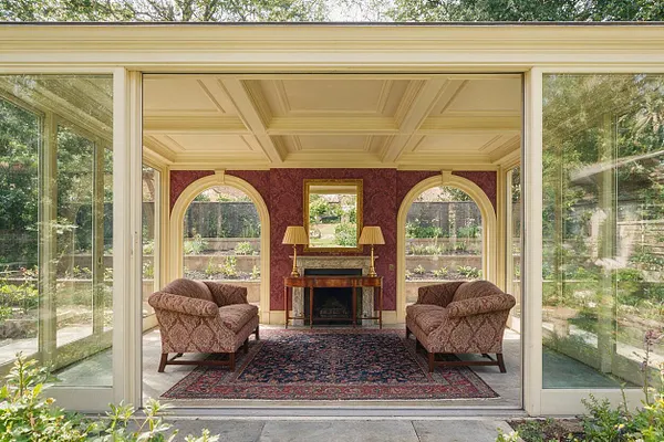 A traditional backyard garden room enclosed by glass walls, featuring a coffered ceiling, arched side openings, and a centered fireplace with a mirror above it. Two patterned upholstered chairs face inward on a dark oriental rug, creating a symmetrical sitting area with views to the landscaped garden outside.