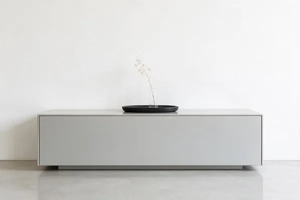 A sparse minimalist interior with a low, rectangular light gray cabinet centered against a plain white wall. On top sits a shallow black tray holding a clear glass vessel with a single delicate dried stem, creating a simple sculptural focal point.