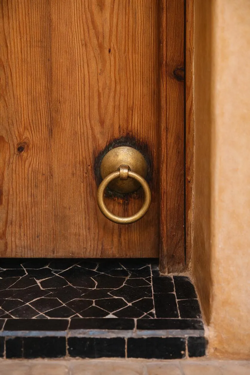 A close-up detail of a rustic wooden door with a round brass ring pull mounted at the center. The door is framed by warm plaster walls and sits above a black stone threshold with visible mosaic-like grout lines.